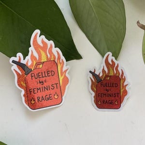 Two red glitter stickers shaped like flaming fuel cans, both reading “FUELLED BY FEMINIST RAGE,” placed on a white surface with large green leaves surrounding them. The stickers have different sizes and both have a white edge.