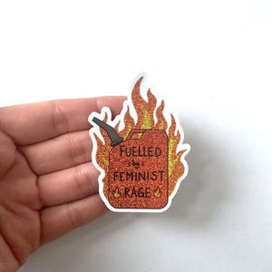 A hand holds a red glitter sticker shaped like a flaming fuel can, reading “FUELLED BY FEMINIST RAGE,” against a plain white background. The sticker has a sparkling finish and a white edge.