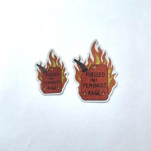 Two red glitter stickers shaped like flaming fuel cans, both reading “FUELLED BY FEMINIST RAGE,” displayed side by side on a plain white background, with one sticker larger than the other. Both sticker have a white edge.