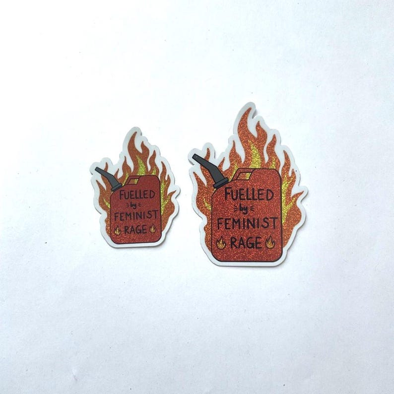 Two red glitter stickers shaped like flaming fuel cans, both reading “FUELLED BY FEMINIST RAGE,” displayed side by side on a plain white background, with one sticker larger than the other. Both sticker have a white edge.