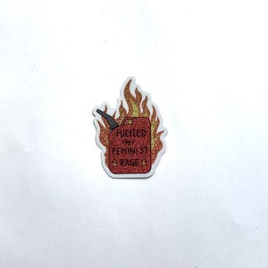A red glitter sticker shaped like a flaming fuel can with yellow and orange flames, reading “FUELLED BY FEMINIST RAGE,” centered on a plain white background.