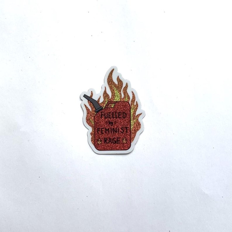 A red glitter sticker shaped like a flaming fuel can with yellow and orange flames, reading “FUELLED BY FEMINIST RAGE,” centered on a plain white background.