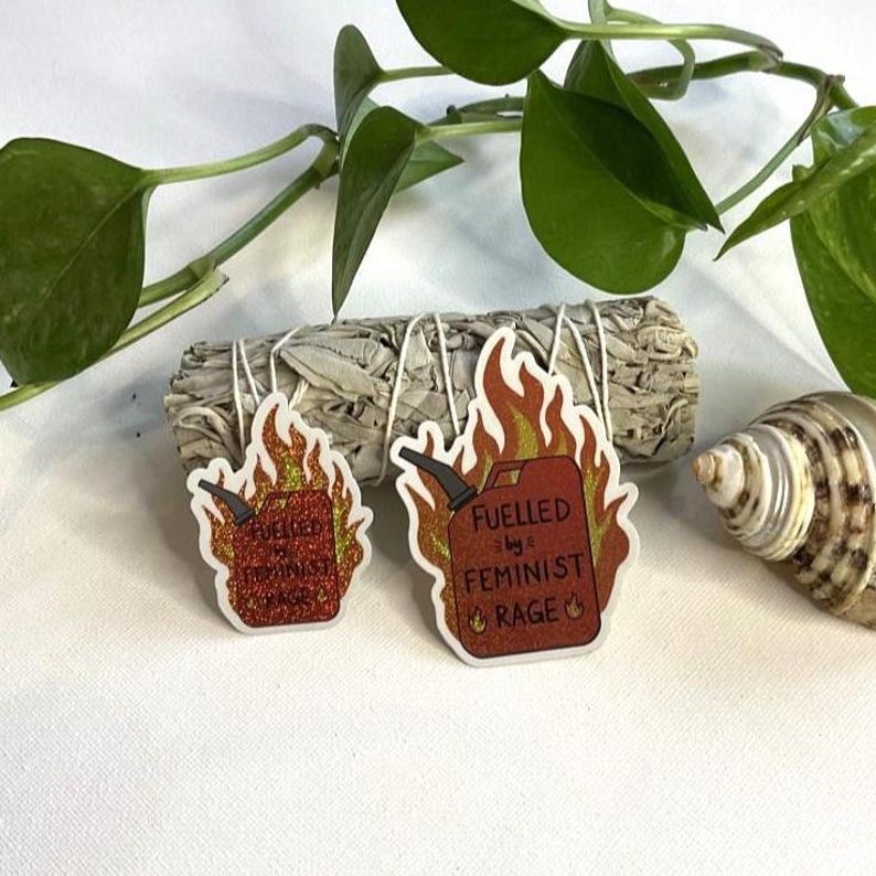 Two red glitter fuel-can stickers reading “FUELLED BY FEMINIST RAGE” arranged on white fabric beside a seashell and bundled sage, with trailing green plant leaves framing the scene.