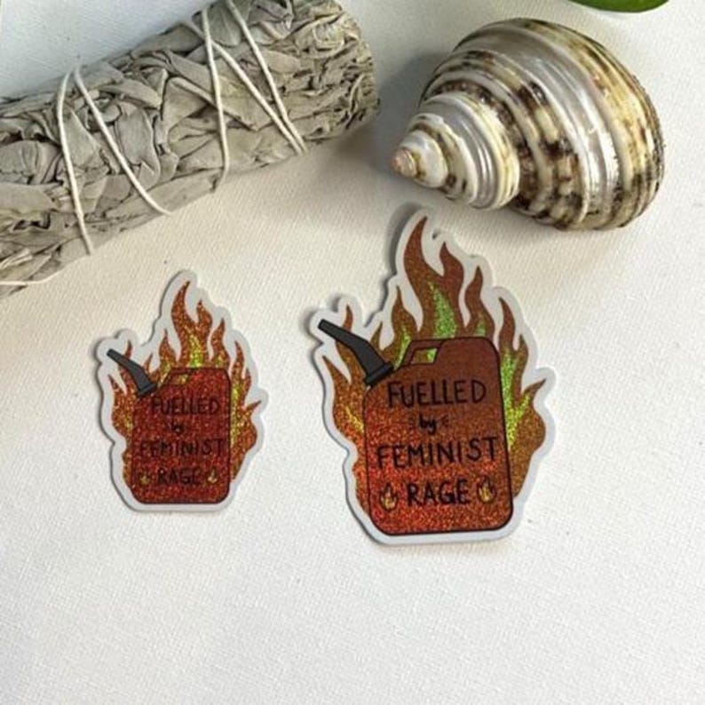 Two red glitter stickers shaped like flaming fuel cans, reading “FUELLED BY FEMINIST RAGE,” arranged on white ground beside a seashell, a bundled sage stick, and trailing green plant leaves.