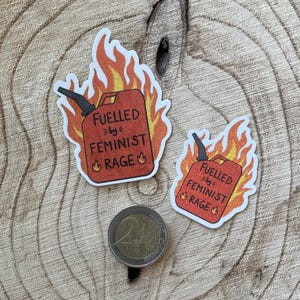 Two red glitter stickers shaped like flaming fuel cans, reading “FUELLED BY FEMINIST RAGE,” displayed on a textured wooden surface with a 2-euro coin beside them for scale.