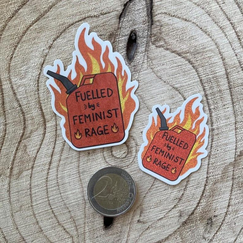 Two red glitter stickers shaped like flaming fuel cans, reading “FUELLED BY FEMINIST RAGE,” displayed on a textured wooden surface with a 2-euro coin beside them for scale.