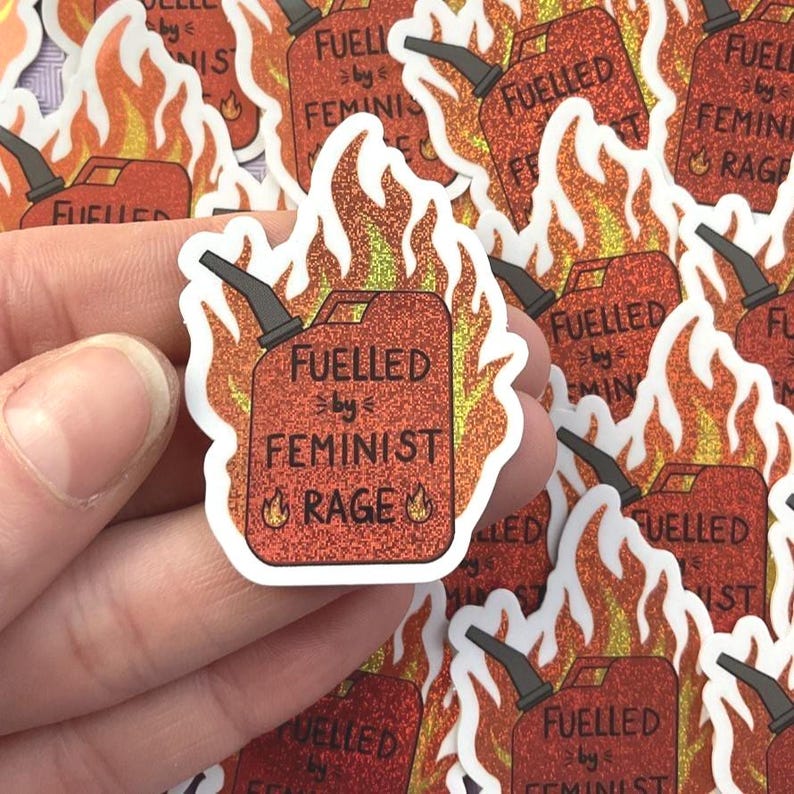 A hand holds a red glitter sticker reading “FUELLED BY FEMINIST RAGE,” designed like a flaming petrol can, with multiple identical stickers arranged on the background behind it.
