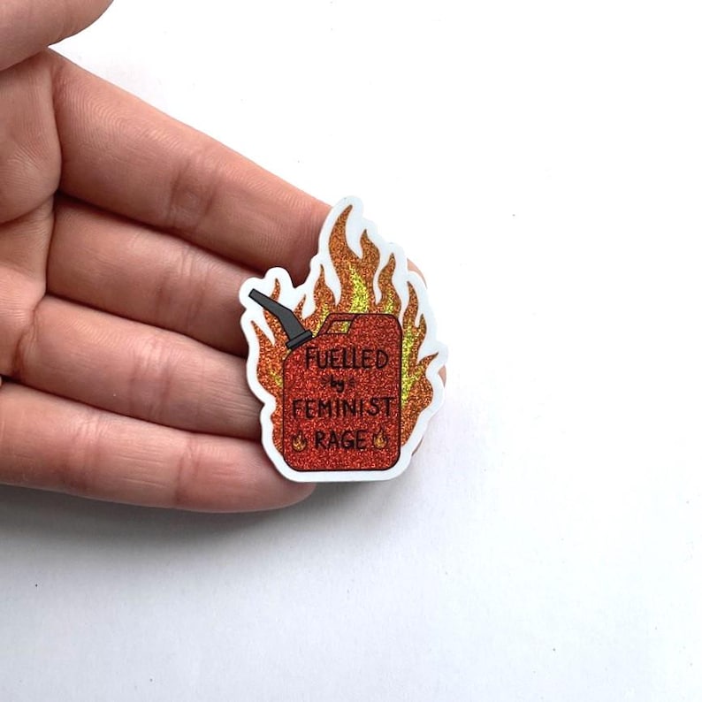 A hand holds a small red glitter sticker shaped like a flaming fuel can with a white edge, reading “FUELLED BY FEMINIST RAGE,” against a plain white background.