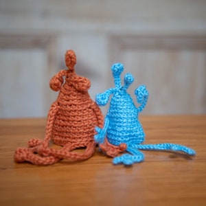 Op de afbeelding: Twee gehaakte amigurumi-speelgoedjes, een oranje en een blauwe, met lange ledematen en unieke kenmerken. De speelgoedjes zitten op een houten oppervlak.