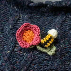 May include: A crocheted flower with a pink and orange center, and a crocheted bee with black and yellow stripes, on a dark blue knitted fabric.