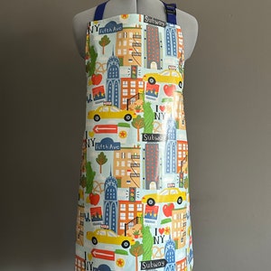 Op de afbeelding: Een waterdichte schort in blauw en wit met een New York City-thema. De schort heeft een herhalend patroon van gele taxi's, gebouwen en de tekst "I Love NY", "Fifth Ave" en "Subway".