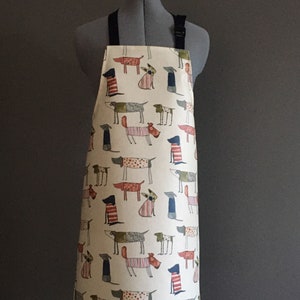 Könnte beinhalten: Eine weiße Schürze mit einem Muster aus bunten Hunden. Die Schürze hat verstellbare Träger und eine Tasche.