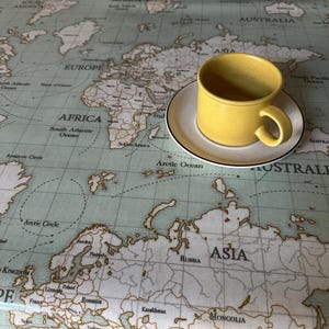 May include: A yellow ceramic coffee cup and saucer set on a tablecloth printed with a world map design. The cup has a curved handle and sits on a white saucer with a black trim. The map is in shades of green and brown.