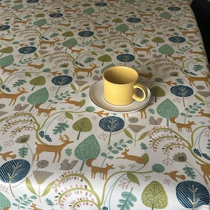 May include: A white tablecloth with a repeating pattern of stylised trees, deer, and birds in shades of green, brown, and yellow. A yellow ceramic mug sits on a white saucer on the tablecloth.