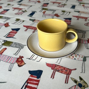 Könnte beinhalten: Eine gelbe Keramik-Kaffeetasse und Untertasse auf einer weißen Tischdecke mit Hundemustern. Die Hunde sind in verschiedenen Farben und Mustern dargestellt. Die Tasse hat eine einfache, zylindrische Form.