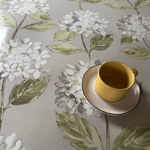 Pode incluir: Uma toalha de mesa cinza claro com um padrão floral de hortênsias em branco, cinza e verde. Uma chávena de chá amarela está sobre um pires branco com uma borda preta. A toalha de mesa é provavelmente de tecido.