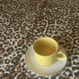 Puede incluir: Una taza de café de cerámica amarilla con un platillo blanco, sentada sobre un mantel de estampado de leopardo marrón y negro.