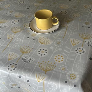 May include: A grey tablecloth with a yellow and white floral pattern. A yellow ceramic mug sits on a white saucer on the tablecloth.