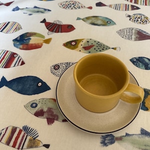 May include: A yellow ceramic coffee cup and saucer with a black rim on a white tablecloth with a pattern of colourful fish.