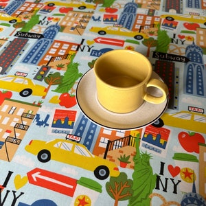 Puede incluir: Una taza de café amarilla con un platillo se encuentra sobre un mantel azul y blanco con temática de la ciudad de Nueva York. El mantel presenta ilustraciones de edificios, taxis y la Estatua de la Libertad. El texto "Subway", "Fifth Ave" e "I NY" es visible en el mantel.