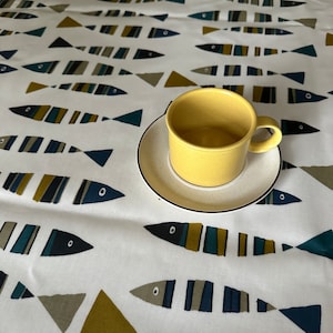 May include: A white tablecloth with a repeating pattern of blue, yellow and green fish. A yellow ceramic mug sits on a white saucer in the centre of the tablecloth.