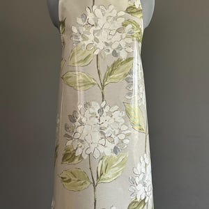 May include: An apron with a floral hydrangea pattern in white, gray, and green on a beige background. The apron has a lime green neck strap and a plastic-like sheen.