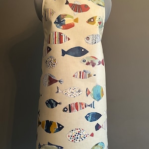 May include: A beige apron with a colorful fish pattern. The fish are various shades of blue, red, yellow, and green. The apron has adjustable straps.