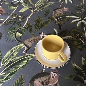 May include: A yellow ceramic teacup and saucer set on a dark grey background with a jungle print. The print features monkeys, palm trees, and large green leaves. The teacup is round with a handle.