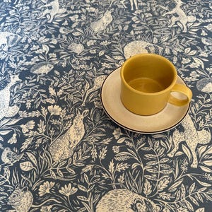 May include: A blue tablecloth with a white woodland pattern featuring animals like deer, rabbits, and a badger. A yellow ceramic mug sits on a white saucer on the tablecloth.
