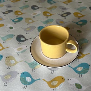 May include: A yellow ceramic teacup and saucer with a black rim on a tablecloth with a repeating pattern of colourful birds.