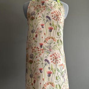 May include: Floral patterned apron with red, purple, and white flowers on a cream backdrop. The apron has a light green neck strap. The apron is displayed on a mannequin.