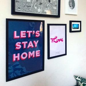 Puede incluir: Una impresi&oacute;n enmarcada con el texto "Let's Stay Home" en letras 3D rosas sobre un fondo azul oscuro. La impresi&oacute;n est&aacute; enmarcada en negro y est&aacute; colgada en una pared blanca.