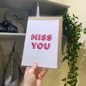 Puede incluir: Una tarjeta blanca con las palabras "MISS YOU" en letras rosas y rojas con lunares blancos. La tarjeta está sostenida en una mano.