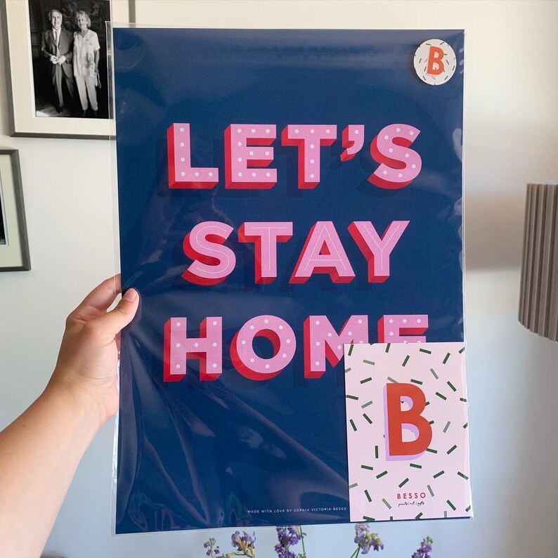 Puede incluir: Un p&oacute;ster azul con el texto "Let's Stay Home" en letras 3D rosas con lunares blancos. El p&oacute;ster est&aacute; enmarcado en una funda de pl&aacute;stico transparente. Una peque&ntilde;a tarjeta rosa con una letra "B" blanca y un patr&oacute;n de confeti se encuentra en la esquina inferior derecha.