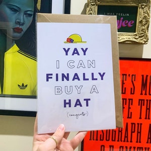 Puede incluir: Una tarjeta blanca con texto morado que dice "YAY I CAN FINALLY BUY A HAT (congrats!)". La tarjeta tiene un sombrero amarillo con flores rojas y azules.