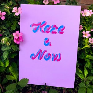 Puede incluir: Un póster rosa con el texto "Here & Now" en letras 3D azules y rosas.