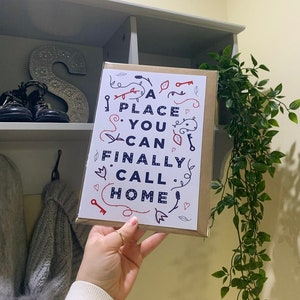 Puede incluir: Una tarjeta blanca con un diseño azul y rojo que dice "A place you can finally call home". La tarjeta está siendo sostenida por una mano con un anillo dorado.