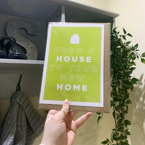 Puede incluir: Una tarjeta verde con texto blanco que dice "From a house to your new home". Un pequeño icono de casa blanca está por encima del texto. La tarjeta está siendo sostenida por una mano.