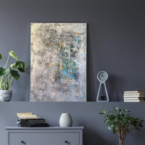May include: Abstract painting with shades of gray, white, and blue. The painting is framed and hanging on a gray wall. There is a clock and a plant on a shelf below the painting.