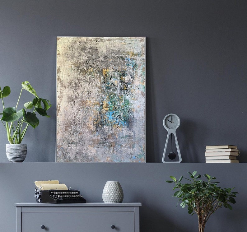 May include: Abstract painting with shades of gray, white, and blue. The painting is framed and hanging on a gray wall. There is a clock and a plant on a shelf below the painting.