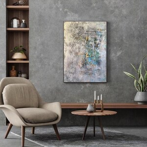 May include: A beige armchair with wooden legs sits in front of a gray wall with a large abstract painting hanging above a wooden shelf. A round wooden table with a silver vase and candlesticks sits in front of the armchair.