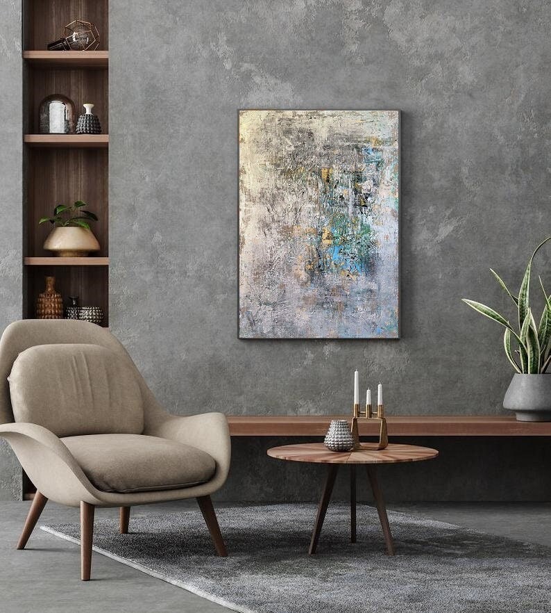 May include: A beige armchair with wooden legs sits in front of a gray wall with a large abstract painting hanging above a wooden shelf. A round wooden table with a silver vase and candlesticks sits in front of the armchair.