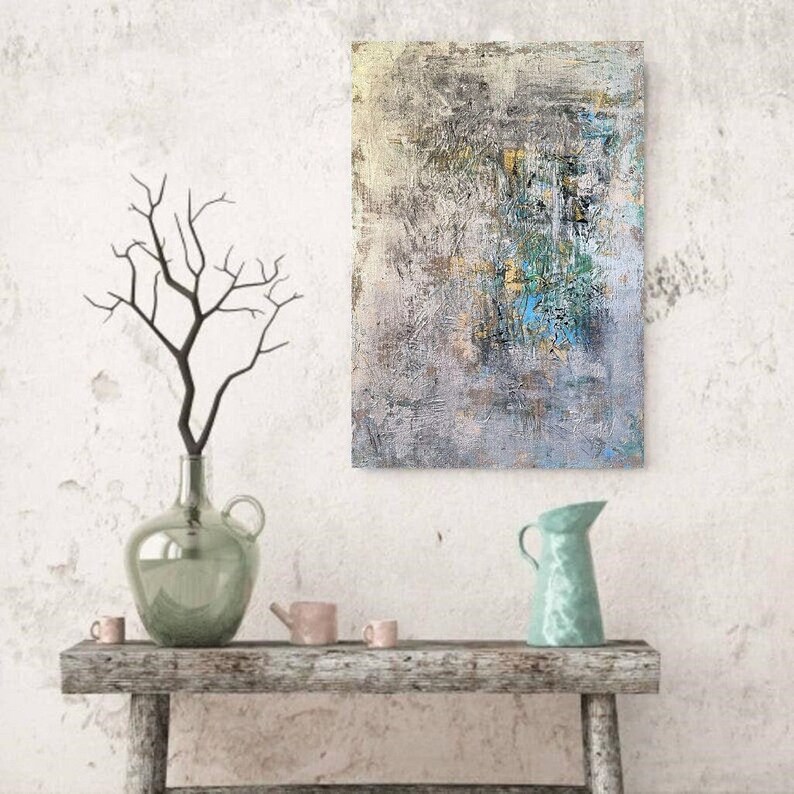May include: Abstract painting with shades of gray, blue, and yellow. The painting is hanging on a white wall above a wooden shelf with a vase, a pitcher, and two small cups.