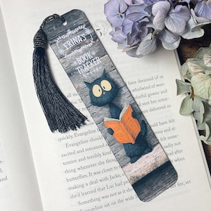 May include: A bookmark with a black tassel, depicting a cartoon black cat reading an orange book. The bookmark has the text "ERINA'S BOOK TRACKER" on a wood-grain background. The bookmark is on an open book.