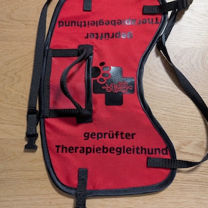 Könnte beinhalten: Eine rote Hunde-Weste mit schwarzen Verzierungen und Riemen. Auf der Weste steht in Weiß "geprüfter Therapiebegleithund", zusammen mit einem Pfotenabdruck und einem Kreuz-Logo. Sie hat verstellbare Riemen und eine Tasche.