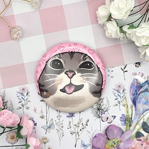 Puede incluir: Una insignia redonda con la cara de un gato gris con una capucha rosa adornada con corazones y estrellas. El gato tiene una expresión alegre. El fondo incluye cuadros rosas, papel floral y flores artificiales.