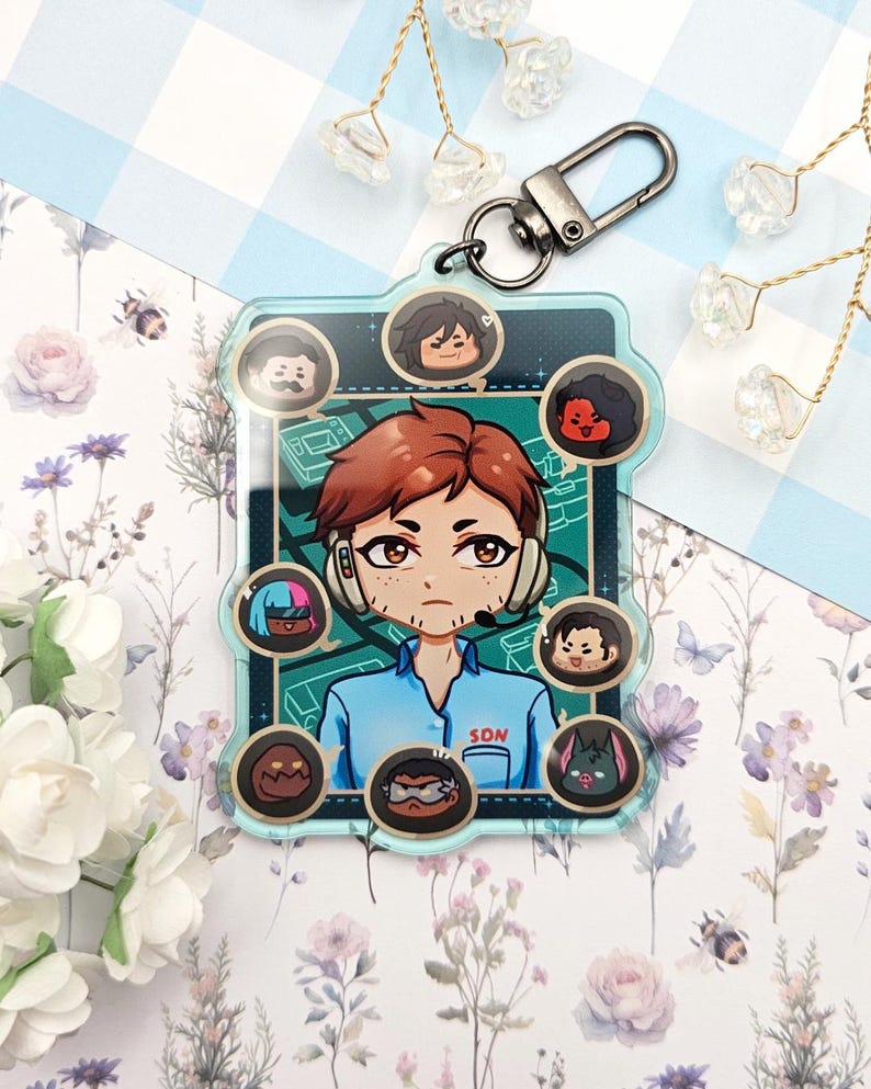 Pode incluir: Um chaveiro de acr&iacute;lico transparente com uma ilustra&ccedil;&atilde;o de desenho animado de uma pessoa com fones de ouvido e o texto "SON". O chaveiro tem um fecho prateado e inclui v&aacute;rios pequenos retratos de personagens.