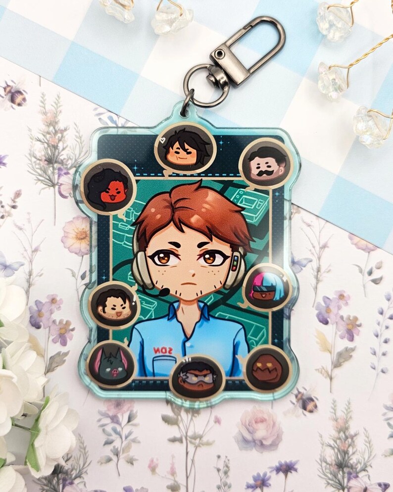 Pode incluir: Um chaveiro de acr&iacute;lico transparente com um personagem de desenho animado com cabelo ruivo e um fone de ouvido, rodeado por retratos de personagens menores. O chaveiro tem um fecho prateado e o texto "MOR" &eacute; vis&iacute;vel na camisa do personagem. O fundo &eacute; um padr&atilde;o floral.