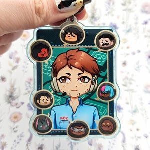 Pode incluir: Um chaveiro de acr&iacute;lico transparente com um personagem central de desenho animado com cabelo ruivo e um fone de ouvido, cercado por retratos de personagens menores. O chaveiro tem um fecho prateado e um fundo verde-azulado com o texto "MO2".