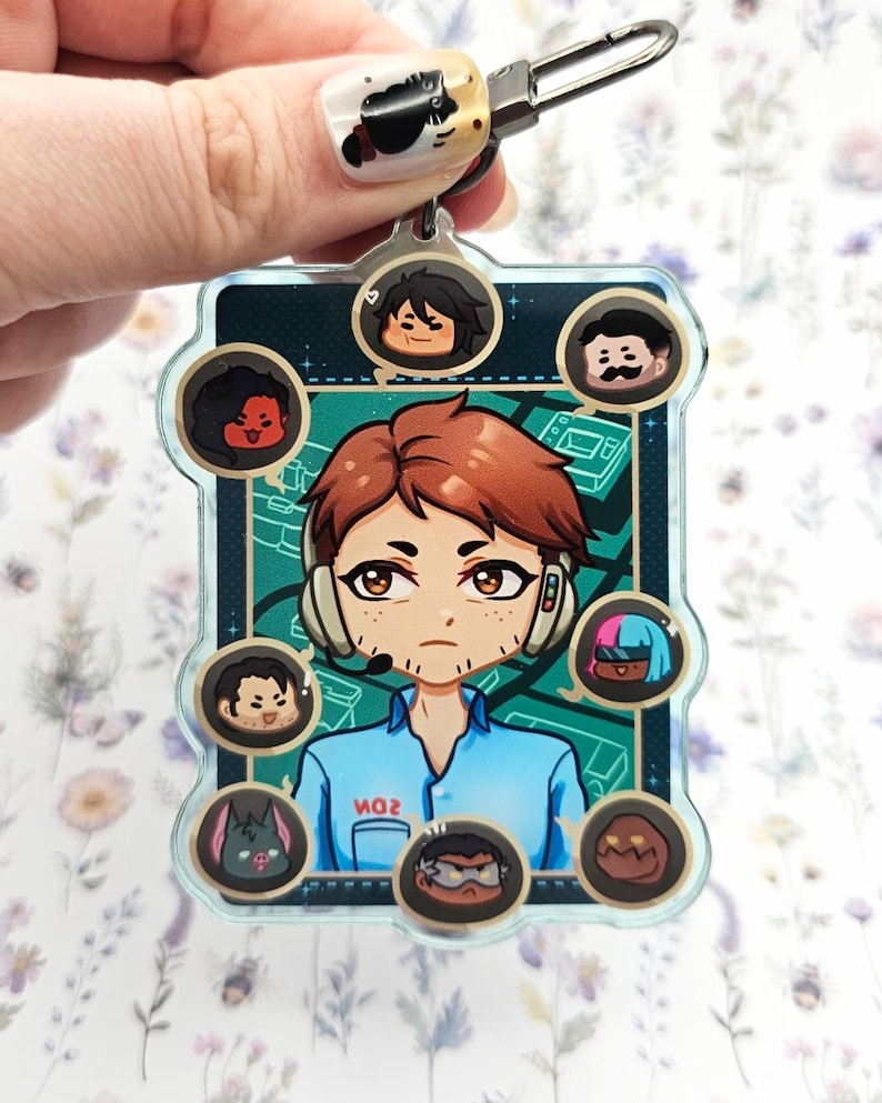 Pode incluir: Um chaveiro de acr&iacute;lico transparente com um personagem central de desenho animado com cabelo ruivo e um fone de ouvido, cercado por retratos de personagens menores. O chaveiro tem um fecho prateado e um fundo verde-azulado com o texto "MO2".